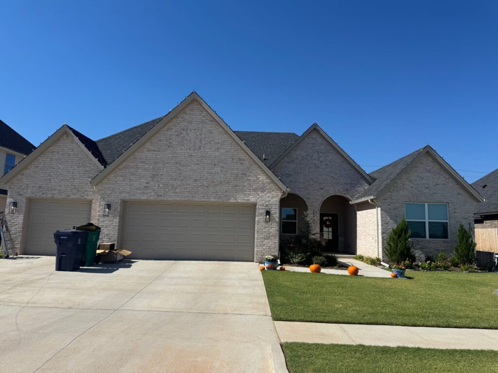 A brick home in Yukon/Piedmont, OK is outlined along the roofline with multicolor nostalgic Christmas lights, with a neat lawn, driveway, and a few pumpkins decorating the front walkway