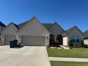 A brick home in Yukon/Piedmont, OK is outlined along the roofline with multicolor nostalgic Christmas lights, with a neat lawn, driveway, and a few pumpkins decorating the front walkway